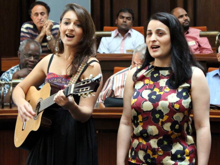 Zwei Frauen stehen in der Mitte des Hodlersaals und singen. Eine davon spielt auf einer akustischen Gitarre, die sie in der Hand hält.